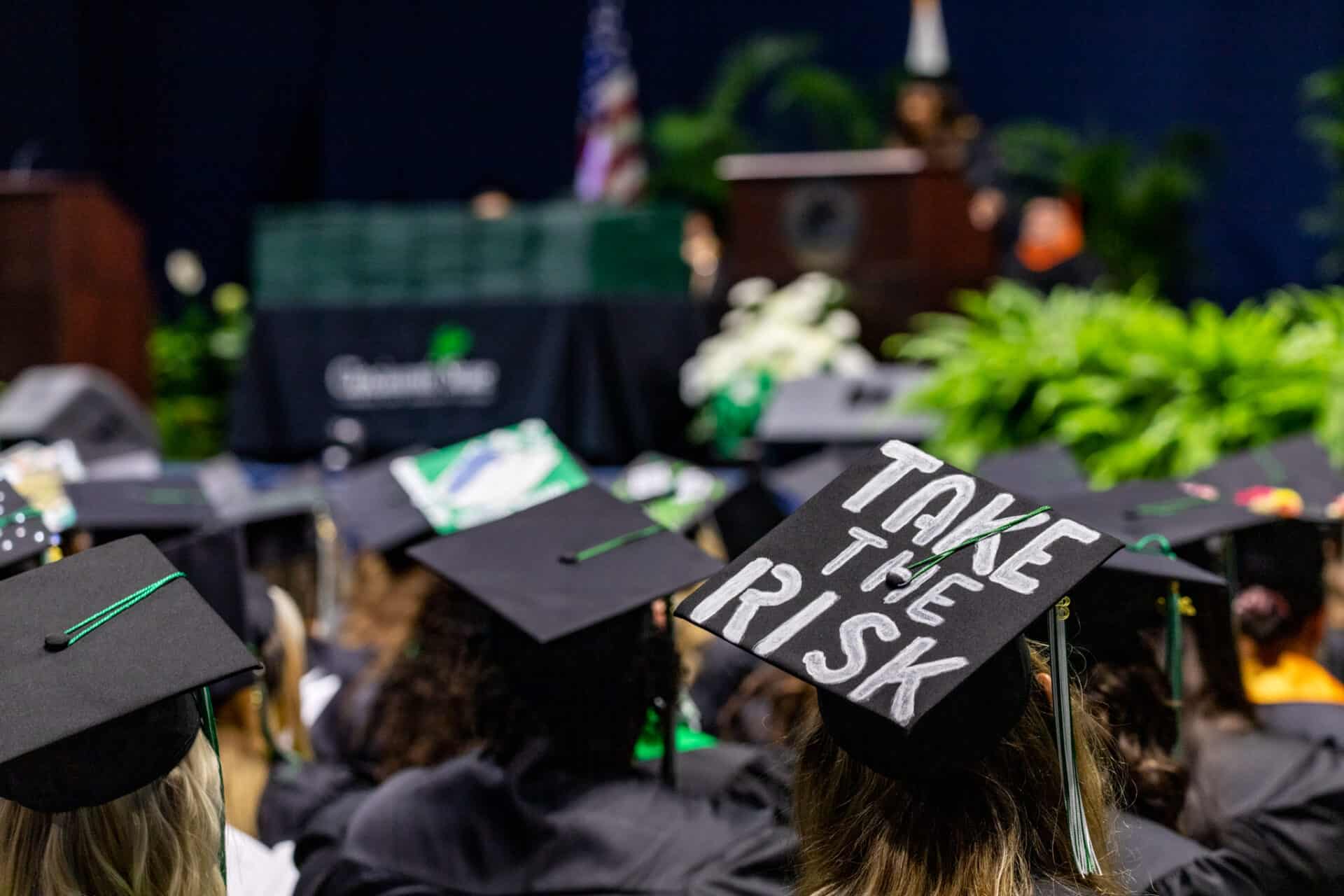 group of graduation caps in front of the commencement stage with one cap reading Take The Risk